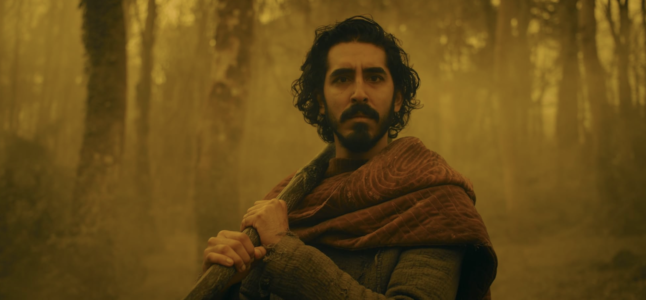 Dev Patel as Gawain standing in a forest in yellow mist in The Green Knight (2021) (IMDb)